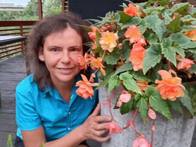 GrostFlow mit lockigem, braunem Haar, einem blauen Hemd und einem Lächeln, hält einen Topf mit orangefarbenen und rosa Blumen direkt vor ihrem Gesicht. Ihre Brüste sind nicht sichtbar..