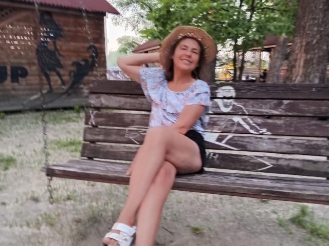 GrostFlow sitting on a wooden bench, wearing a straw hat, light blue floral shirt, black shorts, and white sandals, smiling with one hand behind her head, showing her ass.