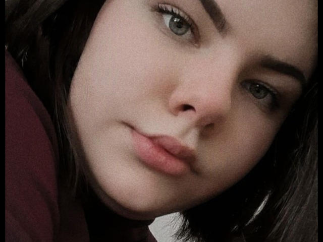 Tinas Moores Gesicht mit blasser Haut, grünen Augen, dunkelbrauen Augenbrauen und vollen, rosafarbenen Lippen. Sie blickt leicht nach rechts und trägt ein dezentes, selbstbewusstes Lächeln..