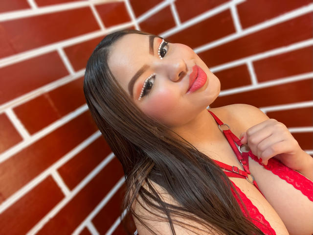 SamanthaFerrer with long brown hair, wearing red lace lingerie, pouting lips, holding strap, exposing large tits, against red brick wall. Her makeup is bold, eyeliner sparkling.