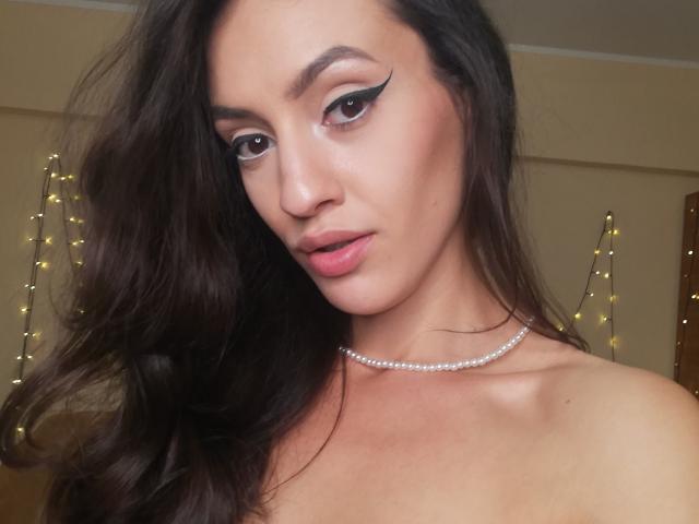 VievienneClemente has long brown hair, dramatic black eyeliner, pink lips, and a pearl necklace, looking seductive with bare shoulders and a soft, confident expression.