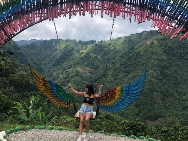 Samantha Morgan mit bunten Flügeln, sitzend auf einer Schaukel, trägt ein schwarzes Oberteil und Jeansshorts, mit Blick auf eine grüne Berglandschaft, unter einem rosa und blauen gewebten Baldachin..
