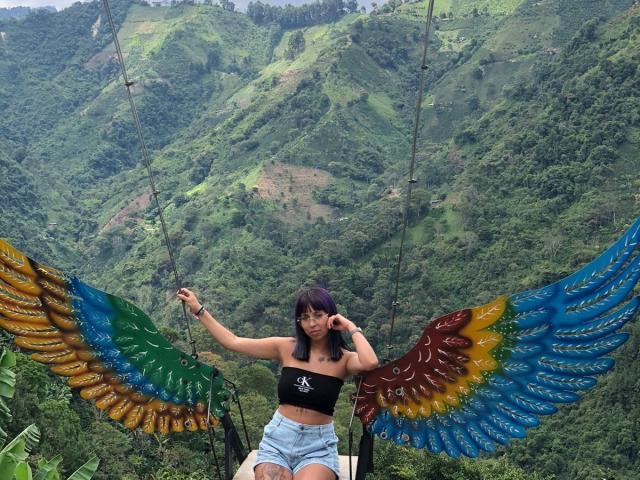 Samantha Morgan, com seios pequenos, vestindo uma cropped preta e shorts azuis, sentada em um balanço com asas coloridas pintadas, em uma área verde e montanhosa, segurando uma corda..