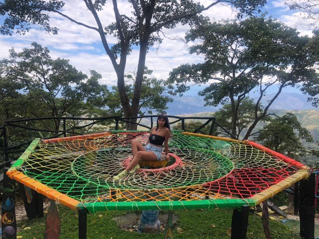 Samantha Morgan, con un trasero grande y pechos pequeños, sentada sobre una red de pesca de colores, vistiendo una camiseta negra y pantalones cortos vaqueros, en una plataforma de madera con árboles y un cielo azul de fondo..