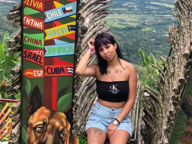 SamanthaMorgan with purple hair, black tube top, showing tits, and denim shorts, tattoo on thigh, standing next to colorful sign, surrounded by cacti. Mountain view in background.