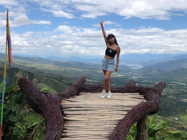 Samantha Morgan se tient sur un pont en bois avec des bords en corde, et pointe vers le haut avec un bras. Elle porte un haut noir à bretelles et un short en jean, avec des baskets blanches..