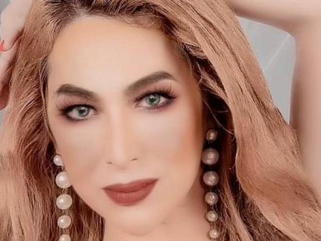 Una donna affascinante, con capelli rosa pallido, occhi verdi e orecchini di perle, ha labbra piene con rossetto rosso scuro e un'espressione seria. La sua mano destra tocca la sua testa..