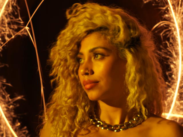 LilithWalker has curly blonde hair, a gold necklace, and a sexy, pensive look with orange lighting. Her tits are slightly visible, and she's looking to the side.