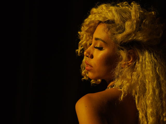 LilithWalker with curly blonde hair, closed eyes, golden glow, bare shoulders, and subtle makeup, showing off her cute nose ring and a small, shiny necklace. Dark background.