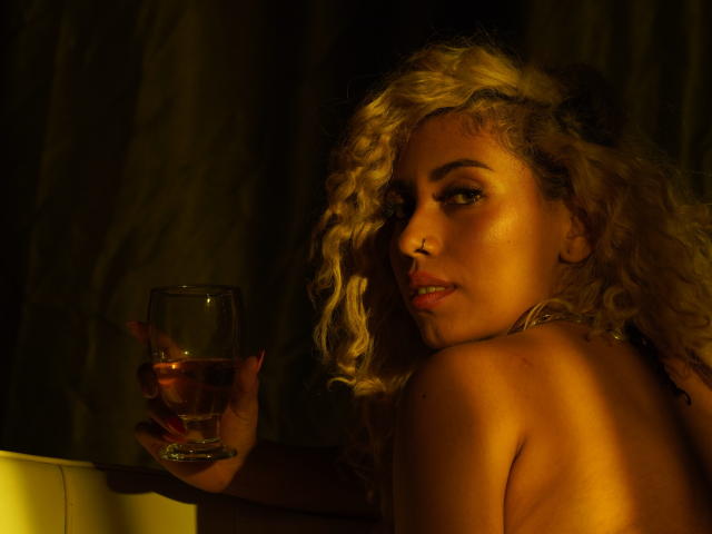 LilithWalker, avec ses cheveux blonds bouclés, tenant un verre de vin rosé, regardant par-dessus son épaule, avec un piercing au nez, une peau éclatante et un sourire séduisant, nue, sur un fond sombre..