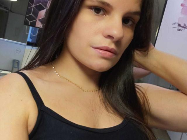 CrisMS with long dark hair, wearing a black tank top, gold necklace, and a serious expression, has small tits and a neutral face, with one arm raised.