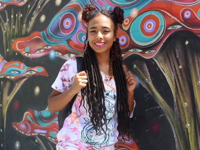 Courtney, met lange zwarte dreadlocks, roze lippenstift en een kleurrijke tie-dye shirt, glimlacht naar de camera. Ze heeft een medium bruine huid en haar borsten zijn klein..