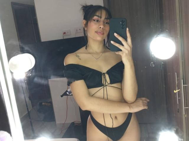 JessicaSoler with small tits, black off-shoulder top, black thong, tattoo on right shoulder, holding phone, mirror selfie, round mirror lights, dark brown hair, light skin, subtle smile.