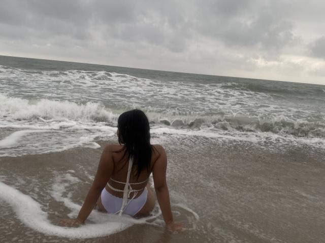 Valery, con cabello negro, bikini blanco y glúteos visibles, sentada en la arena mojada, mirando al océano, con las manos en la espuma blanca, cielo nublado..