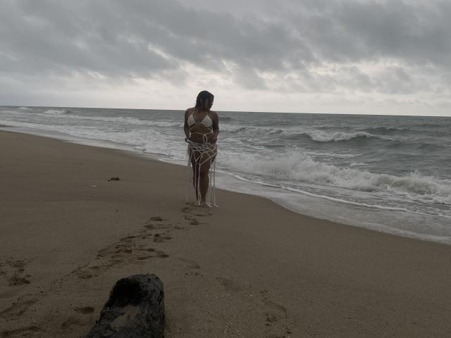 ValerySquirting se tient sur une plage nuageuse, vêtue d'un bikini blanc, les bras serrés contre sa poitrine, avec du sable mouillé et de petites vagues au premier plan..