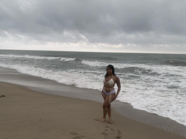 ValerySquirting stands on a sandy beach, wearing a white bikini with straps, her curvy ass and small tits visible, under a cloudy sky with waves in the background.