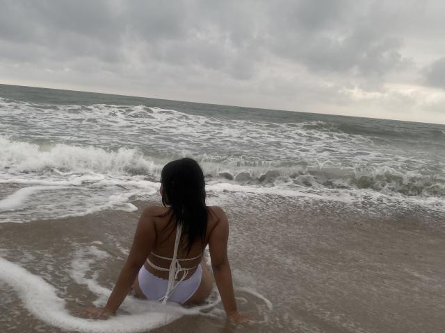 Valery, com cabelos escuros, sentada em uma praia de areia, vestindo um biquíni branco, de costas para a câmera, com os braços na espuma, ondas do oceano, céu nublado. bumbum, seios, biquíni, oceano..