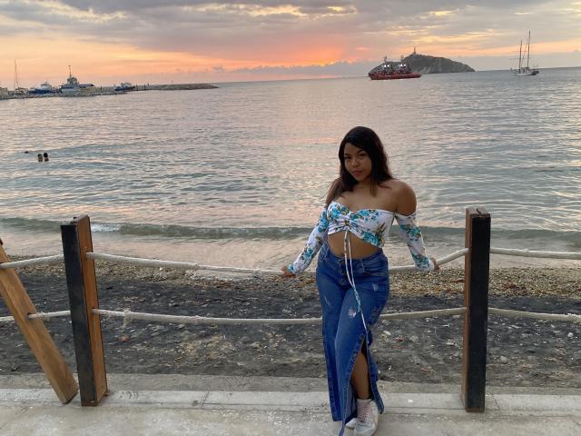 ValerySquirting staat bij het water, gekleed in een blauw-witte bloemen top met een open schouder, blauwe jeans en witte sneakers, met een touwhal in de voorgrond..