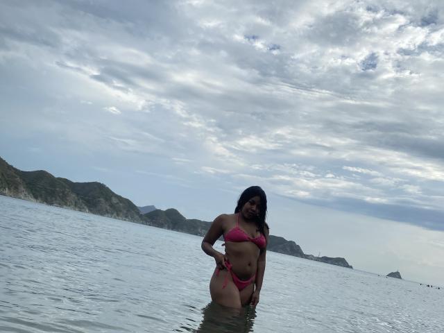 ValerySquirting se tient dans une eau peu profonde, vêtue d'un bikini rose, montrant ses fesses et son vagin, avec un ciel nuageux et des collines lointaines en arrière-plan. Ses cheveux noirs sont mouillés..