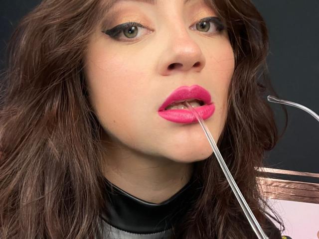 ArleenHunter's face with pink lipstick, brown wavy hair, green eyes, black eyeliner, metal clip in mouth, wearing black and grey leather jacket, serious expression.