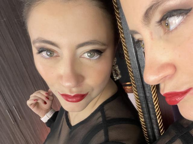 ArleenHunter has green eyes, red lipstick, and is wearing a black mesh top with visible cleavage. Her reflection shows her nose and red lips. She's holding her hair.