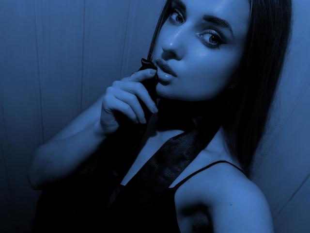 KariMillie with blue-tinted skin, long dark hair, and seductive eyes, biting a black ribbon while wearing a black halter top, showing her small tits and slim arms.