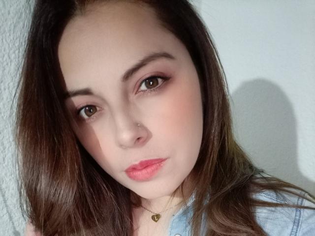 Seher has pale skin, brown eyes, and pink lipstick. She has long brown hair, a nose piercing, and a blue shirt with a heart necklace. Her ass is not visible.