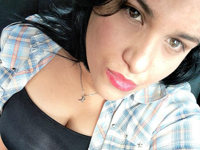 VivianaThompson has pink lips, brown eyes, and black hair, wearing a plaid shirt over a black top, showing her cleavage, with a silver necklace.