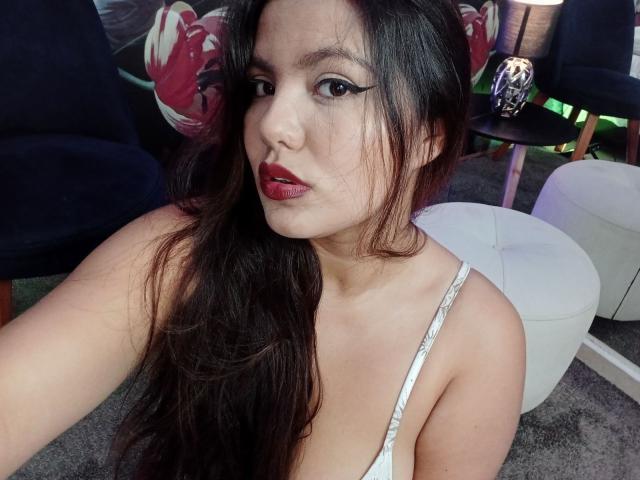 LucyDumont hat langes, dunkles Haar, roten Lippenstift und trägt ein weißes Tanktop, das ihre kleinen Brüste zeigt. Sie sitzt auf einem weißen Hocker..