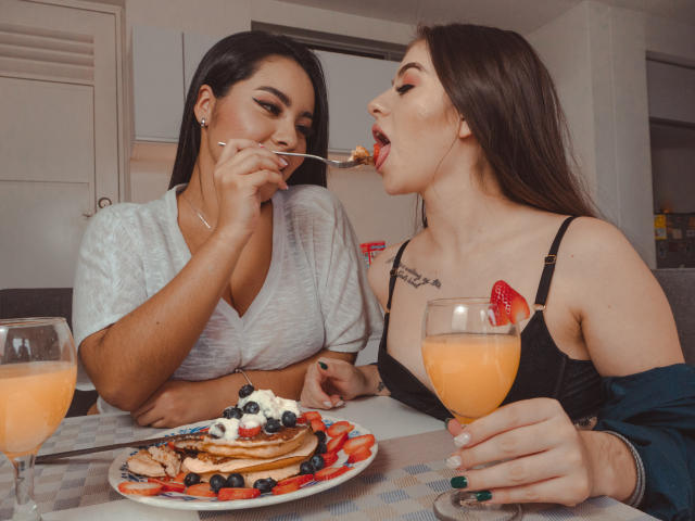 PaolaYKatty with large tits, black hair, eating pancakes with strawberries, blueberries, and cream, drinking orange juice, while another PaolaYKatty with tattoos, black bra, eats from spoon.