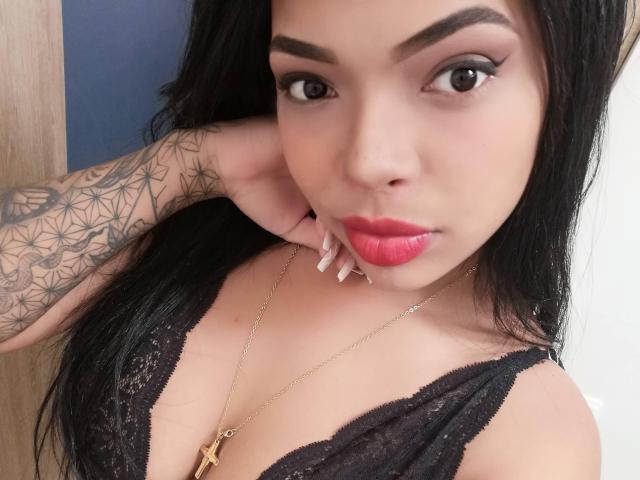 VickyGreys with black lace top, red lipstick, long black hair, tattooed arm, gold cross necklace, pouty lips, and sharp eyeliner, looking at camera, holding her chin.