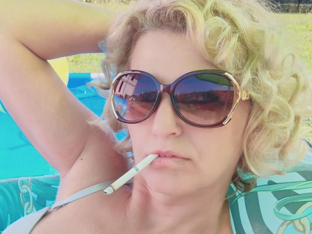 SexyCoco mit lockigem, blondem Haar, die große, braune Sonnenbrillen trägt, hat eine Zigarette im Mund und liegt mit einem Arm hinter dem Kopf..