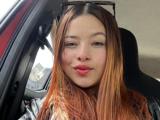 ShantalSummer's face in a car, with long brown hair, blue eyes, pink lips, nose ring, and black sunglasses on her head, smiling slightly.