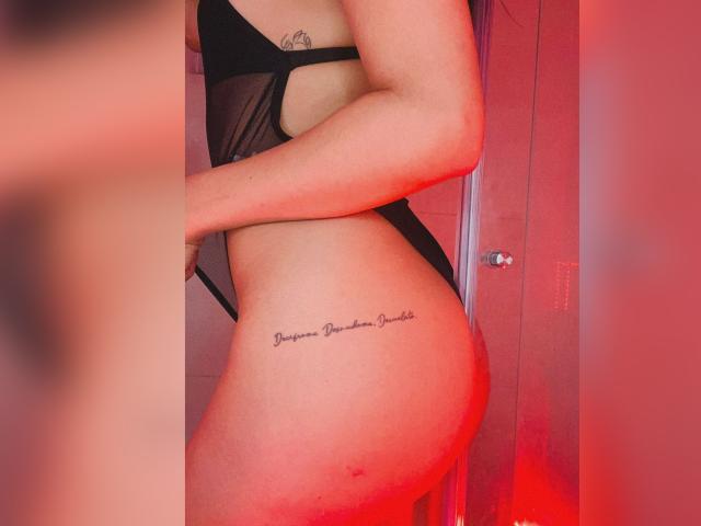 ElizabethMacy in black mesh lingerie, side view, showing her bare ass and a tattoo on her right hip reading "Paparazzi Dreams" under red lighting. Her arm is visible.