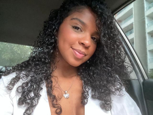 Lisa Mendez, con cabello negro rizado, piel morena y una sonrisa, vestida con una camisa blanca y un collar con forma de corazón, sentada en un coche. Se pueden ver sus senos debajo de la camisa..