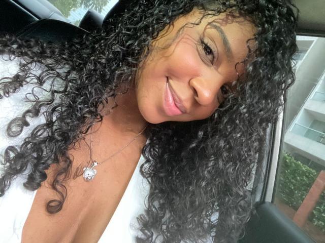 LisaMendez with curly black hair, smiling, wearing a white top, necklace with a flower pendant, showing her large tits, sitting in a car, natural light.