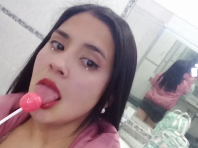 NastyGillyan with dark hair, red lipstick, and pink lollipop in her mouth, looking at the camera, wearing a pink shirt, reflected in a bathroom mirror.