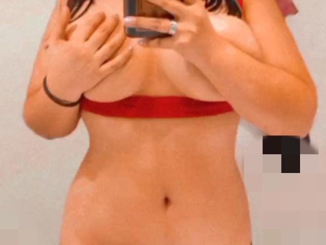 NastyGillyan's tits are covered by a red bra, with her hands holding a phone, showing her bare, tan-skinned, flat-stomached, and nipple-less torso.