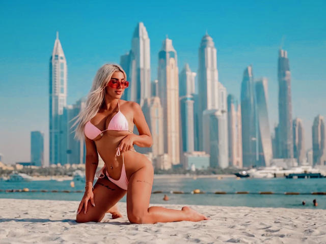 TanyaMonroe with blonde hair, pink bikini, red sunglasses, kneeling on white sand, showing her tits and ass, tattoos on her right thigh, skyscrapers in the background.