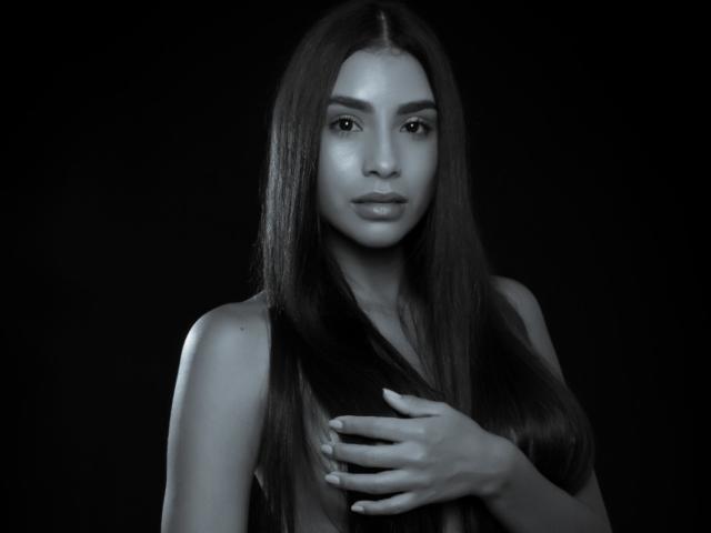 Felicity Adria, mit langem, dunklem Haar, grauer Haut und einem ernsten Gesichtsausdruck, die mit ihrer rechten Hand ihre Brust berührt, trägt ein ärmelloses Oberteil, auf einem Schwarz-Weiß-Foto..