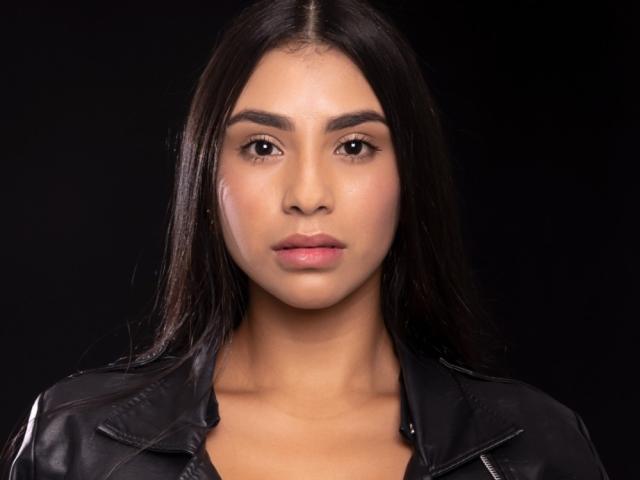 FelicityAdria heeft recht, donkerbruin haar, een medium bruine huidskleur en draagt een zwarte leren jas. Haar gezicht is in het midden geplaatst, met een neutrale uitdrukking, en ze kijkt naar de camera..