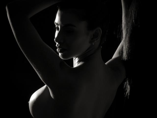 Perfil lateral de FelicityAdria, con sombras marcadas, sin top, con los brazos levantados, mostrando sus pechos, piel lisa, aretes, ligera sonrisa, alto contraste, blanco y negro, sensual, sexy, pose elegante..