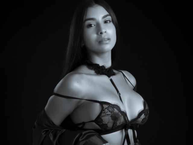 FelicityAdria, met lang donker haar, draagt een zwarte kanten bh en een losvallende robe, waarbij haar borsten zichtbaar zijn, en ze heeft een zelfverzekerde glimlach op haar gezicht op een zwart-wit foto..