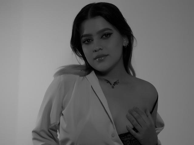 Kim, im Sommer, mit dunklem Haar, trägt ein offenes Hemd und bedeckt ihre Brüste mit einer Hand. Über ihrem Dekolleté ist ein Tattoo zu sehen. Das Foto ist in Schwarzweiß..