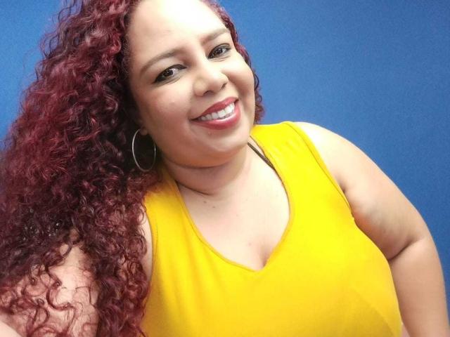 AdelineLox, con i capelli rossi ricci, una maglietta gialla senza maniche, sorridente, che indossa orecchini a cerchio, mostra leggermente il seno, sfondo blu, selfie, pelle media, rossetto rosso..