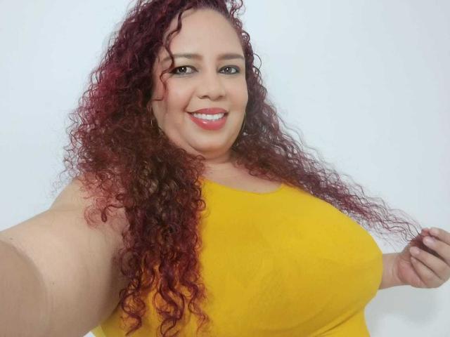 AdelineLox with curly red hair, smiling, wearing a yellow top, showing large boobs, holding her hair, selfie, white background.