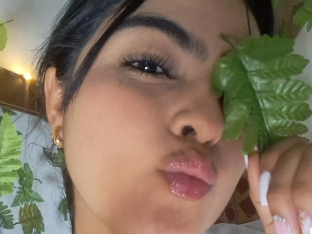 MarilynWoods has a cute face with glossy pink lips, dark eyebrows, and black hair. She's holding green leaves near her cheek with white nails.