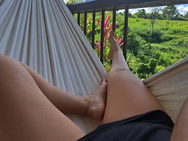 As pernas de Marilyn Woods estão abertas em uma rede bege, mostrando pés descalços e uma pequena saia preta. Seus dedos apontam para cima, com folhagem verde visível do lado de fora..