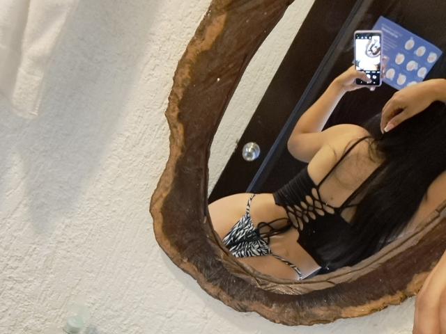 Marilyn Woods em uma selfie no espelho, vestindo uma blusa preta com padrão de treliça e sem costas, e uma parte de baixo com padrão de zebra e estilo fio dental, com cabelos longos pretos, segurando um telefone azul com pontos brancos..