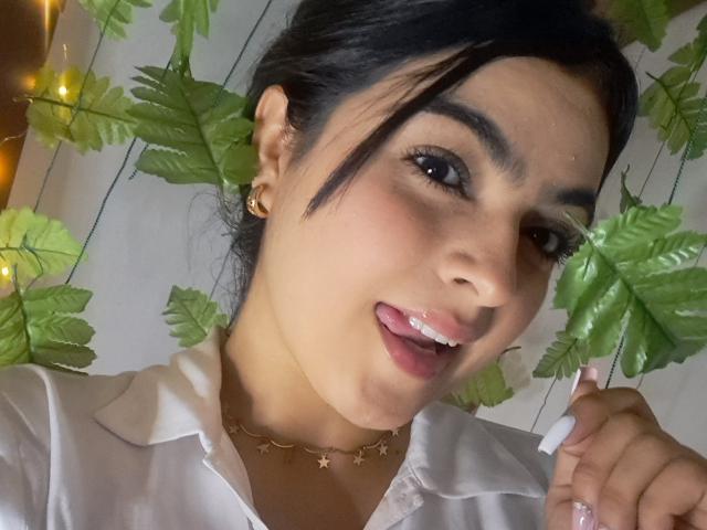 Marilyn Woods, met donker haar, mediumbruine huid en kleine gouden oorbellen, met haar tong uit, een wit shirt, een sterrenketting, een groene, bladerrijke achtergrond en gele lichtjes, met een speelse uitdrukking..
